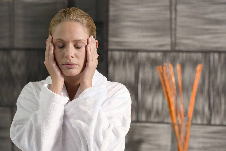 Młoda kobieta w białym szkafroku wykonuje masaż twarzy w eleganckiej łazience