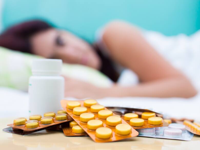 Kobieta leży chora w łóżku, przed nią tabletki na gardło i przeziębienie