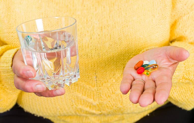 Kobieta w żółtym swetrze trzyma szklankę z wodą i garść tabletek i kapsułek