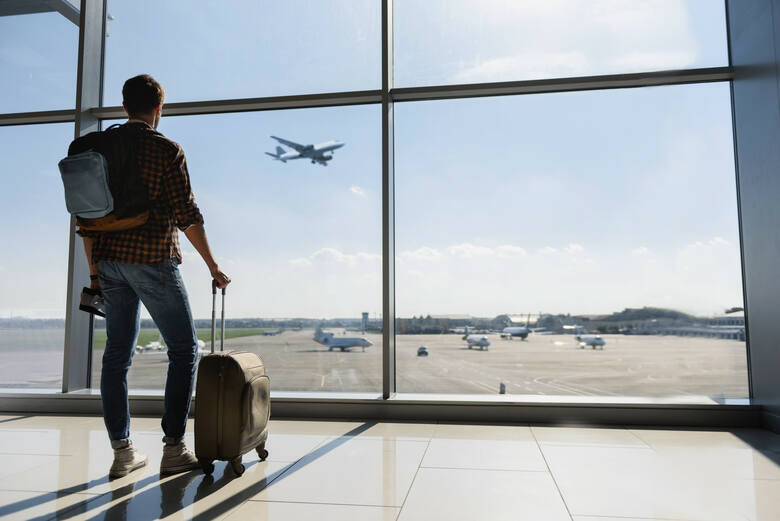 Młody mężczyzna stoi przy oknie na lotnisku i obserwuje samoloty
