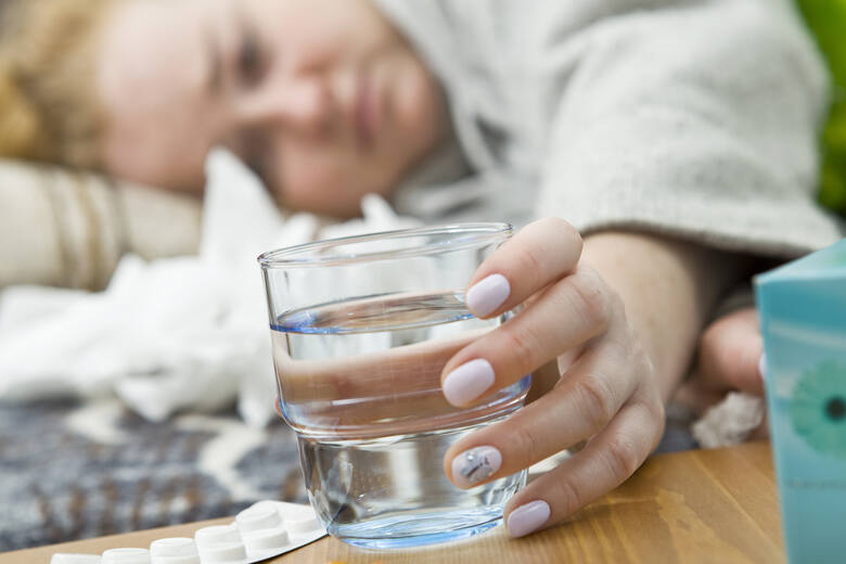 Chora kobieta leży i sięga po szklankę z wodą i leki