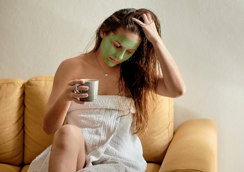 Młoda kobieta w zielonej maseczce kosmetycznej pije z kubka