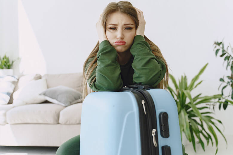 Młoda kobieta z niezadowoloną miną opiera się ramionami o walizkę