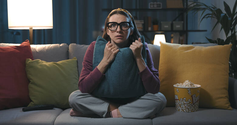 Młoda kobieta w okularach ogląda film, zasłaniając się poduszką