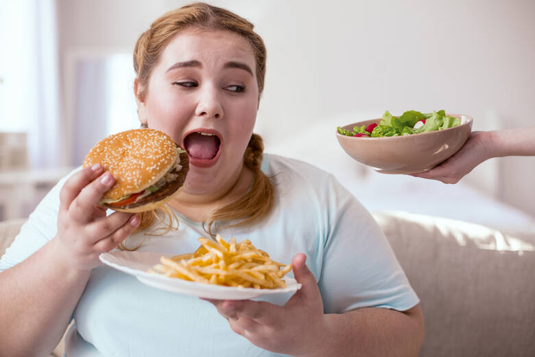 Otyła kobieta je hamburgera i frytki i patrzy na miskę sałatki