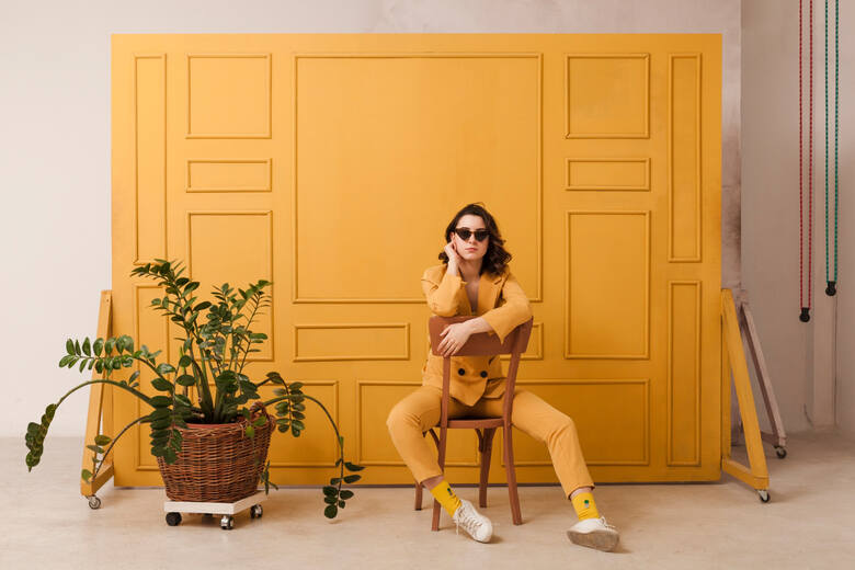 Kobieta w ciemnych okularach i w żółtym ubraniu siedzi na krześle na tle żółtej ściany z kwiatem na podłodze po lewej stronie