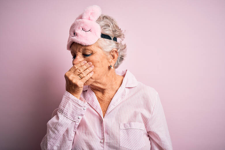 Dojrzała kobieta w różowej piżamie z maseczką do spania na głowie czuje nieprzyjemny zapach