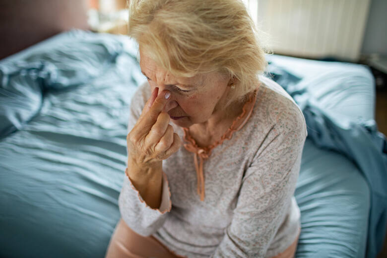Starsza kobieta siedzi na łóżku i uciska nasadę nosa z powodu bólu zatok