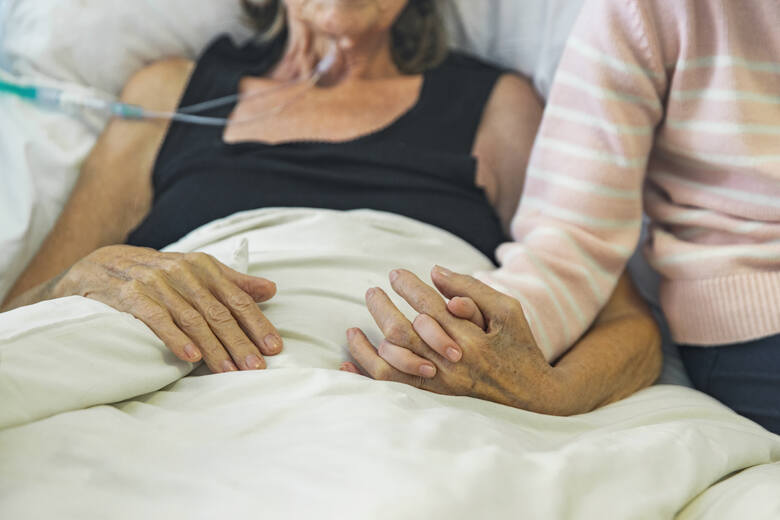 Starsza kobieta w hospicjum trzymana za rękę przez wnuka