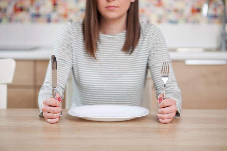 Kobieta siedzi przy stole nad pustym talerzem, w rękach trzyma sztućce