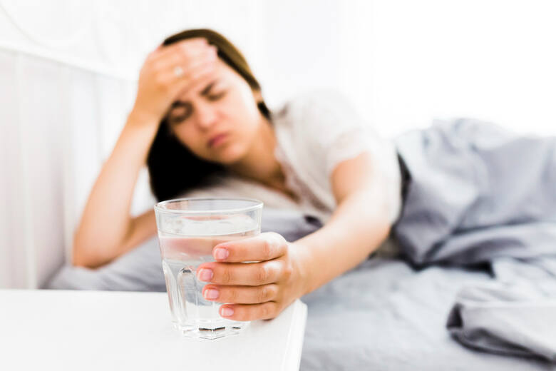 Kobieta z bólem głowy leży w łóżku i sięga po szklankę wody