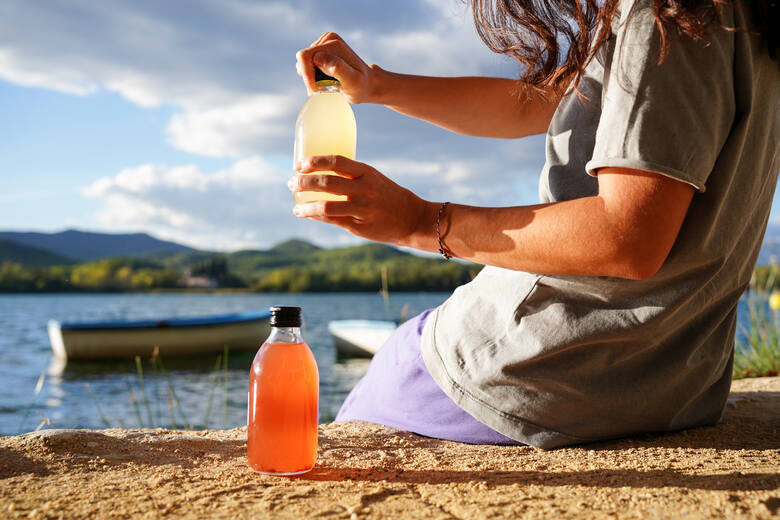 Młoda kobieta siedzi na piasku nad wodą i nalewa kombuchę z butelki