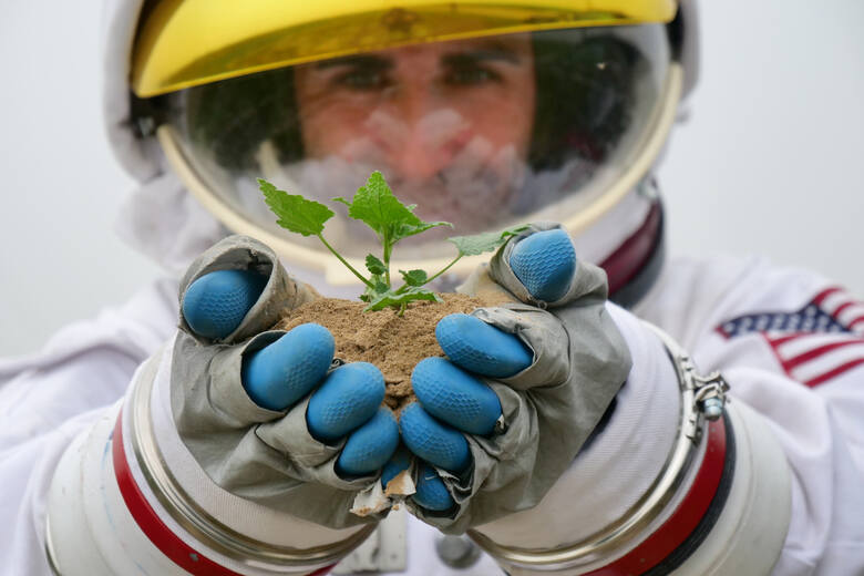 Człowiek w stroju kosmonauty z żywą rośliną i ziemią w dłoniach