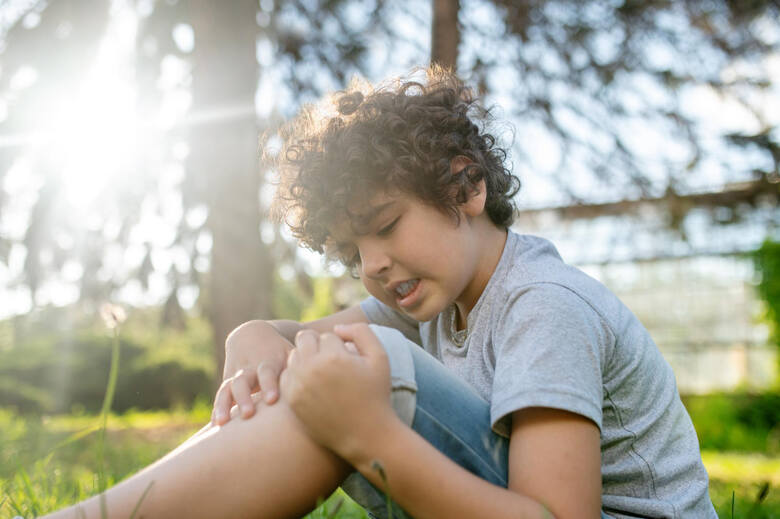 Chłopiec o ciemnych, kręconych włosach siedzi na trawie i drapie kolano