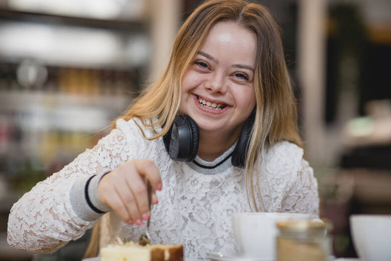 Młoda kobieta z zespołem Downa patrzy w kamerę i śmieje się, siedząc w kawiarni