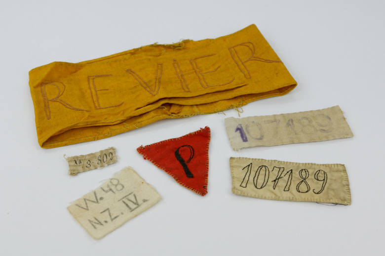 Oznaczenia więźniarskie: numery, czerwony numer noszony przez więźniów politycznych i opaska z napisem po niemiecku „revier”. 