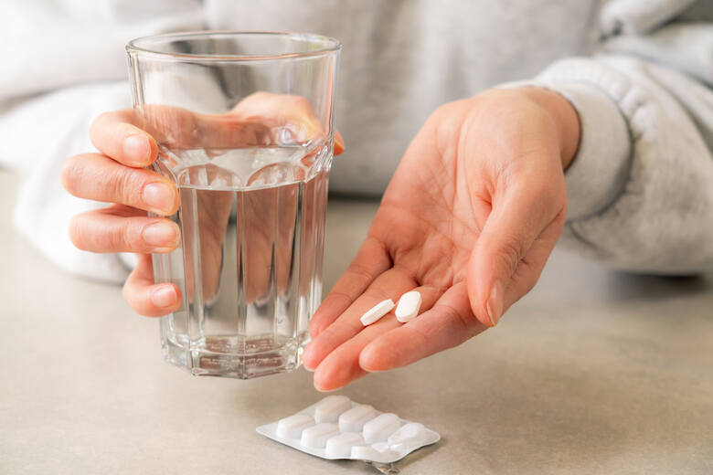 Kobieta trzyma leki przeciwbólowe w jednej ręce i szklankę z wodą w drugiej