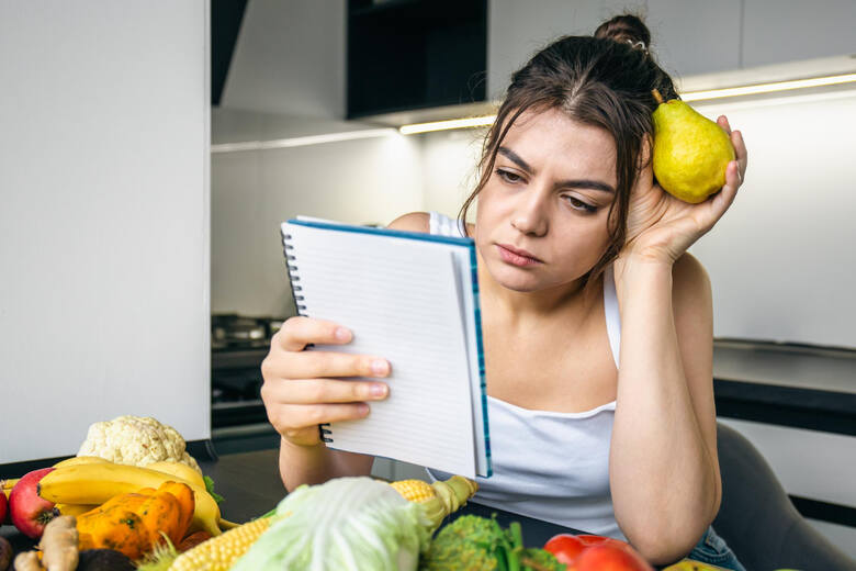 Młoda kobieta z zeszytem i gruszką w dłoni przy stole z owocami i warzywami