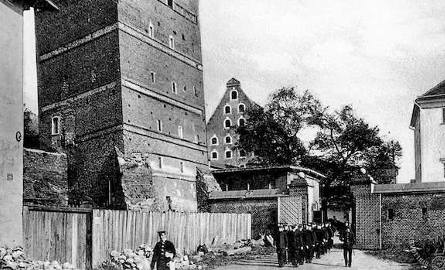 Krzywa Wieża widziana od zewnętrznej strony murów, oraz maszerujący u jej stóp oddział żołnierzy opuszczający koszary racławickie. Tak to wyglądało pod