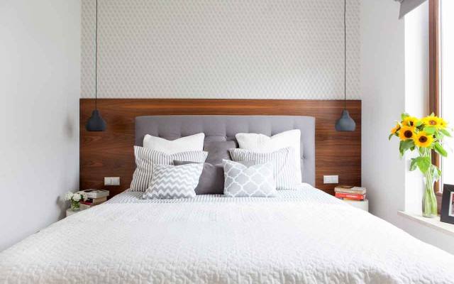 Przytulny nastrój  w tej sypialni tworzy drewno na ścianie za łóżkiem oraz miękkie tekstylia w postaci tapicerowanego zagłówka łóżka i leżących na nim