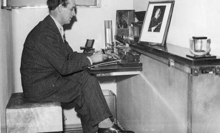 Mistrz fryzjerski Antoni "Antoine" Cierplikowski siedzi przy biurku podczas pracy. Na półce stoją fotografie Benito Mussoliniego i