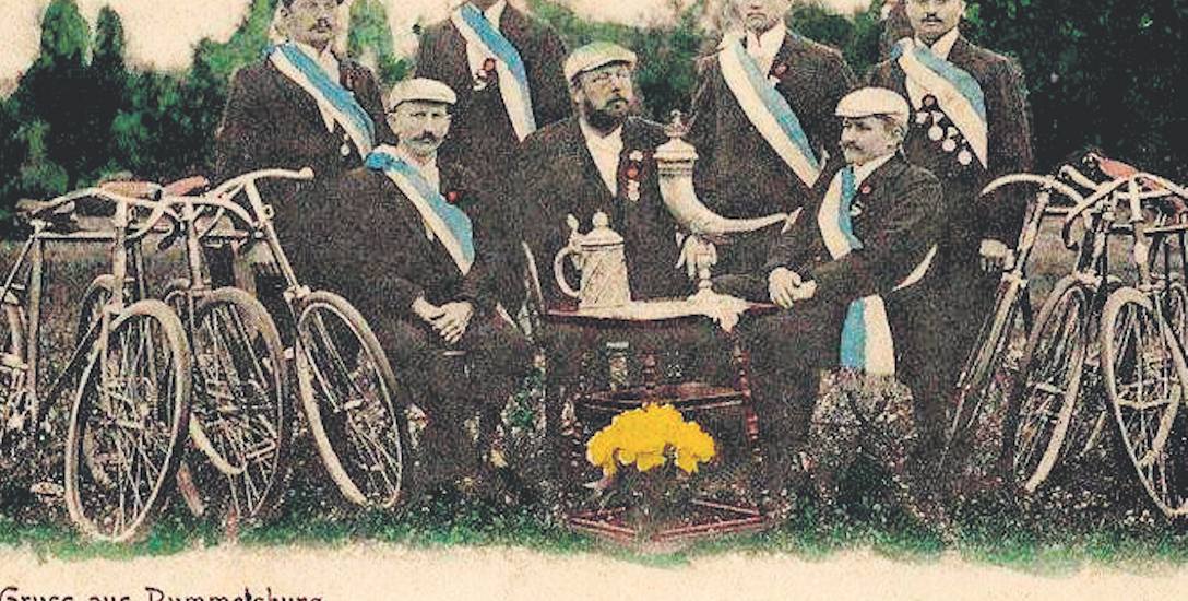 Rowerowe pozdrowienia z Miastka z 1897 roku. Cykliści mają na sobie biało-niebieskie szarfy i medale. Obok stoją ich rowery, w środku stolik m.in. z
