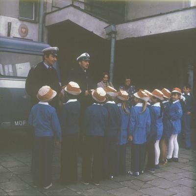 Milicjanci rozmawiają z dziećmi ubranymi w specjalne ostrzegawcze kapelusiki przed budynkiem szkoły. Widoczny fragment samochodu Nysa 522 należacego